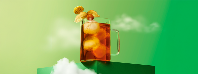 A clear glass filled with richly-coloured iced tea with a ground cherry on the rim, set against a green backdrop with clouds.